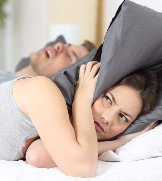 Woman in bed covering her ears with pillow while man snores next to her